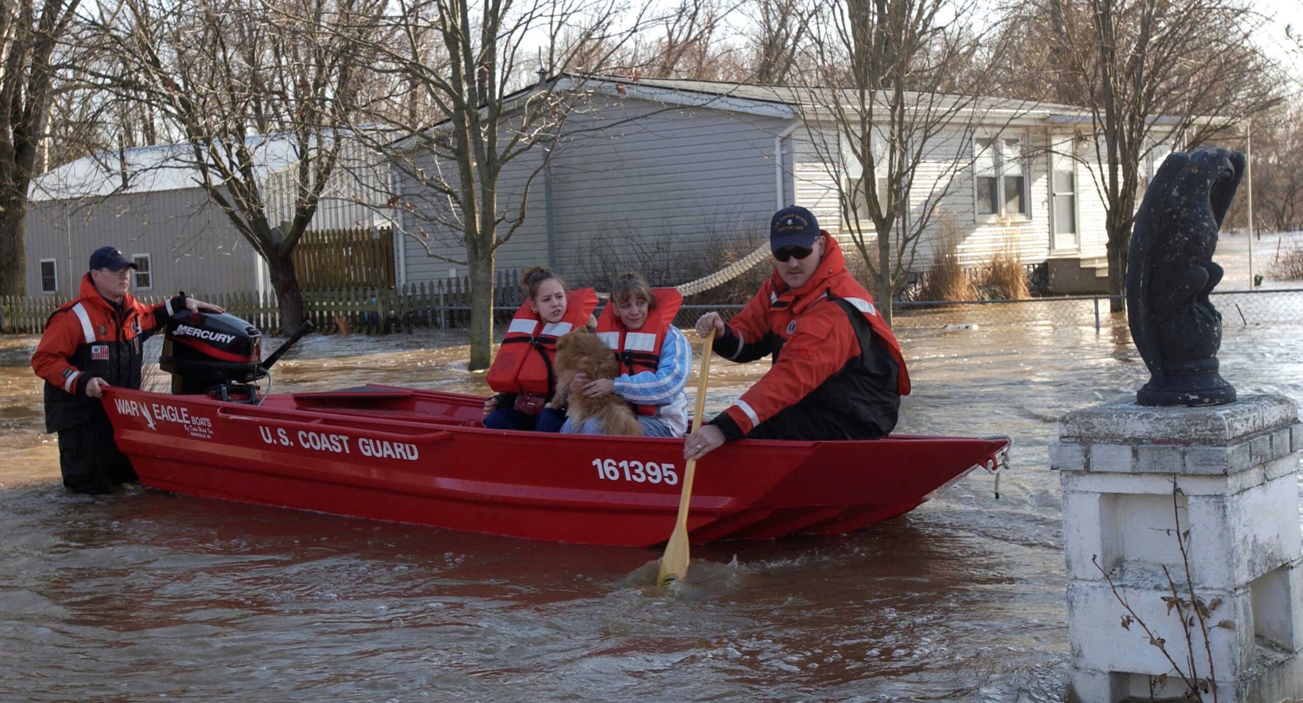 Photo of red U.S. Coast Guard boat in flood water, one paddling, one standing at engine in flood water, with two young girls and dog in boat in residential area.